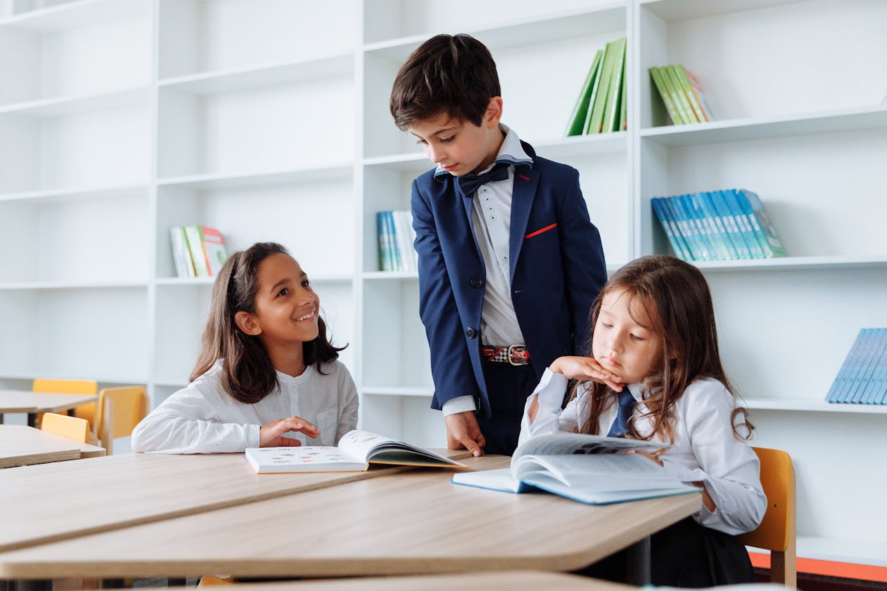 Three children engaging in reading and studying activities at a library table with bookshelves.